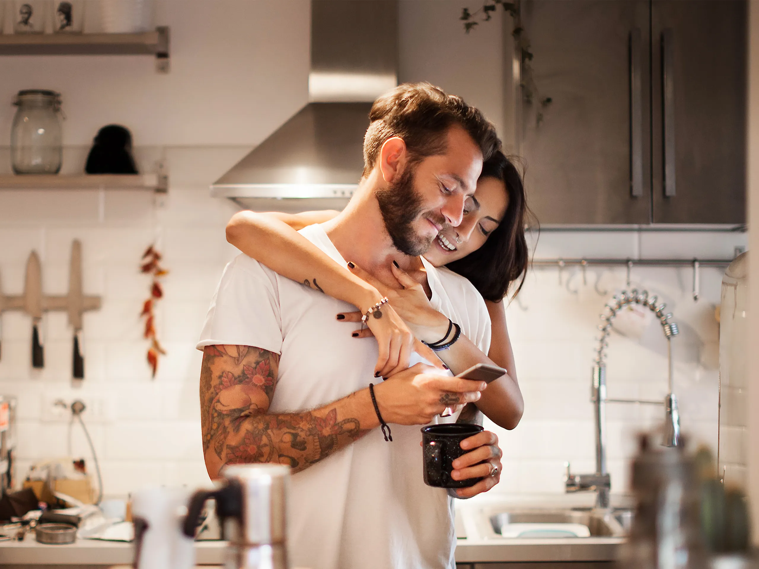 Couple in the kitchen, the man holding a smartphone while the woman hugs him from behind.