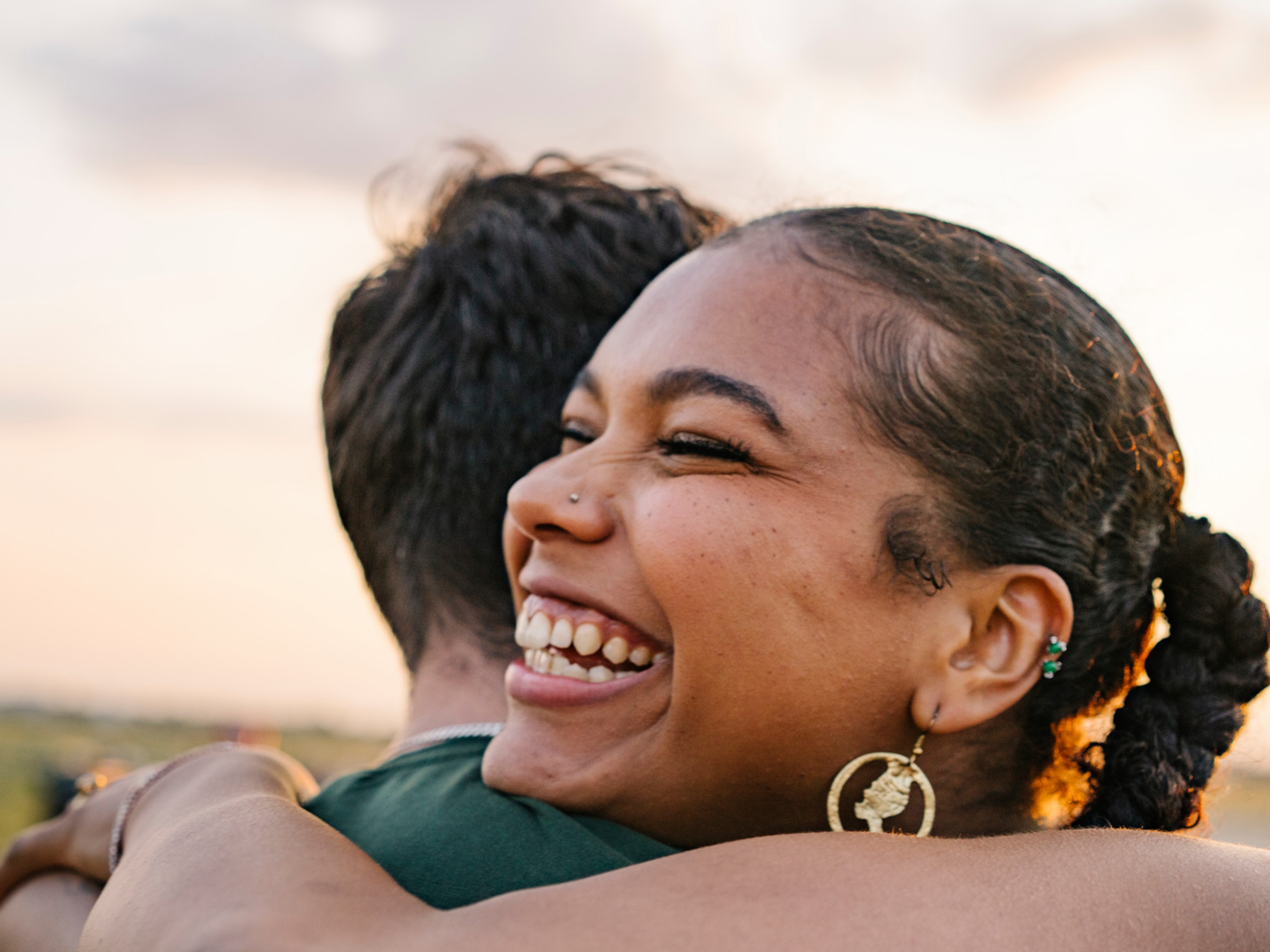 Smiling woman hugging another person outdoors at sunset.