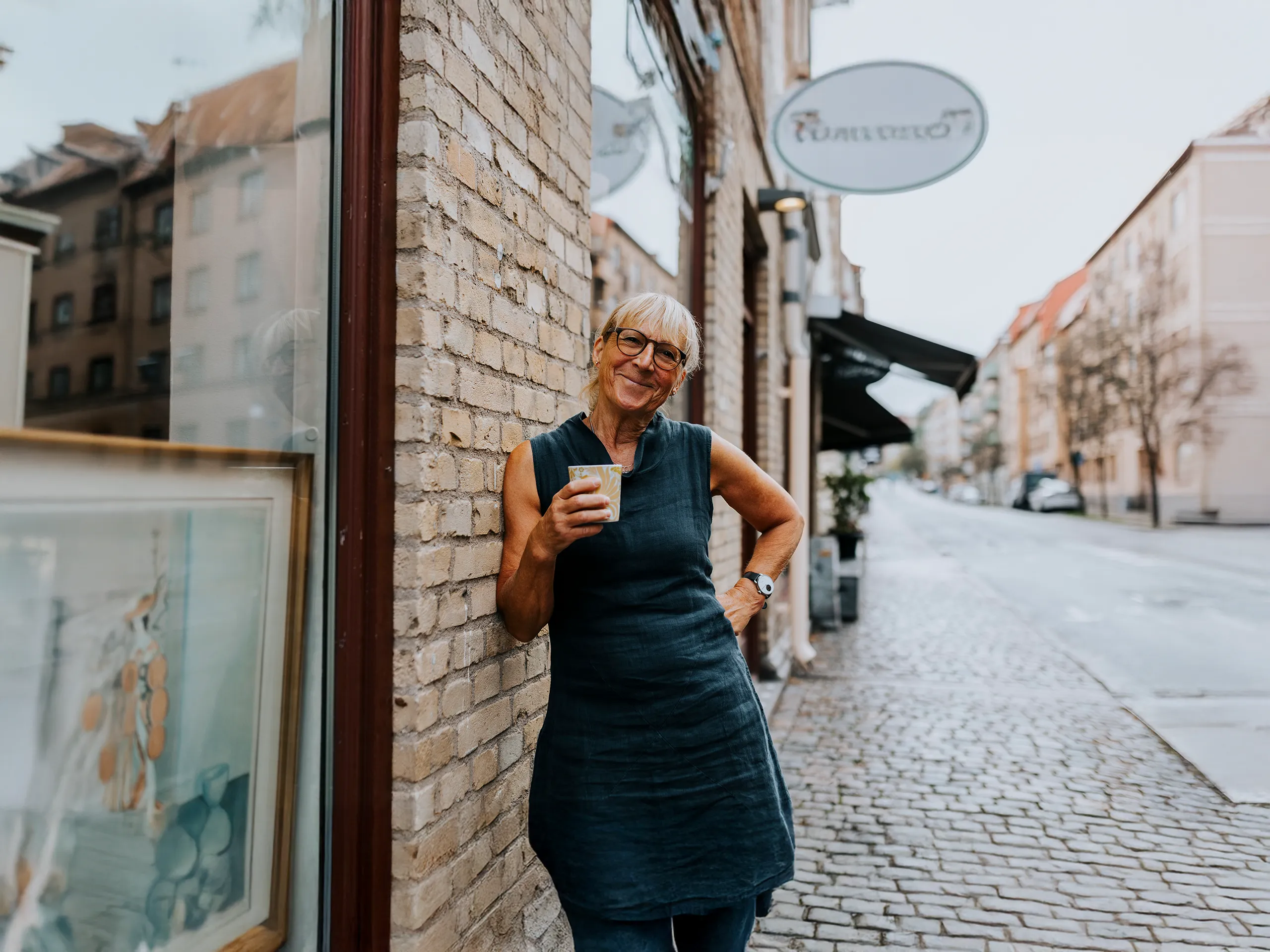 Older woman with coffee leaning and smiling against a house wall on an empty street.
