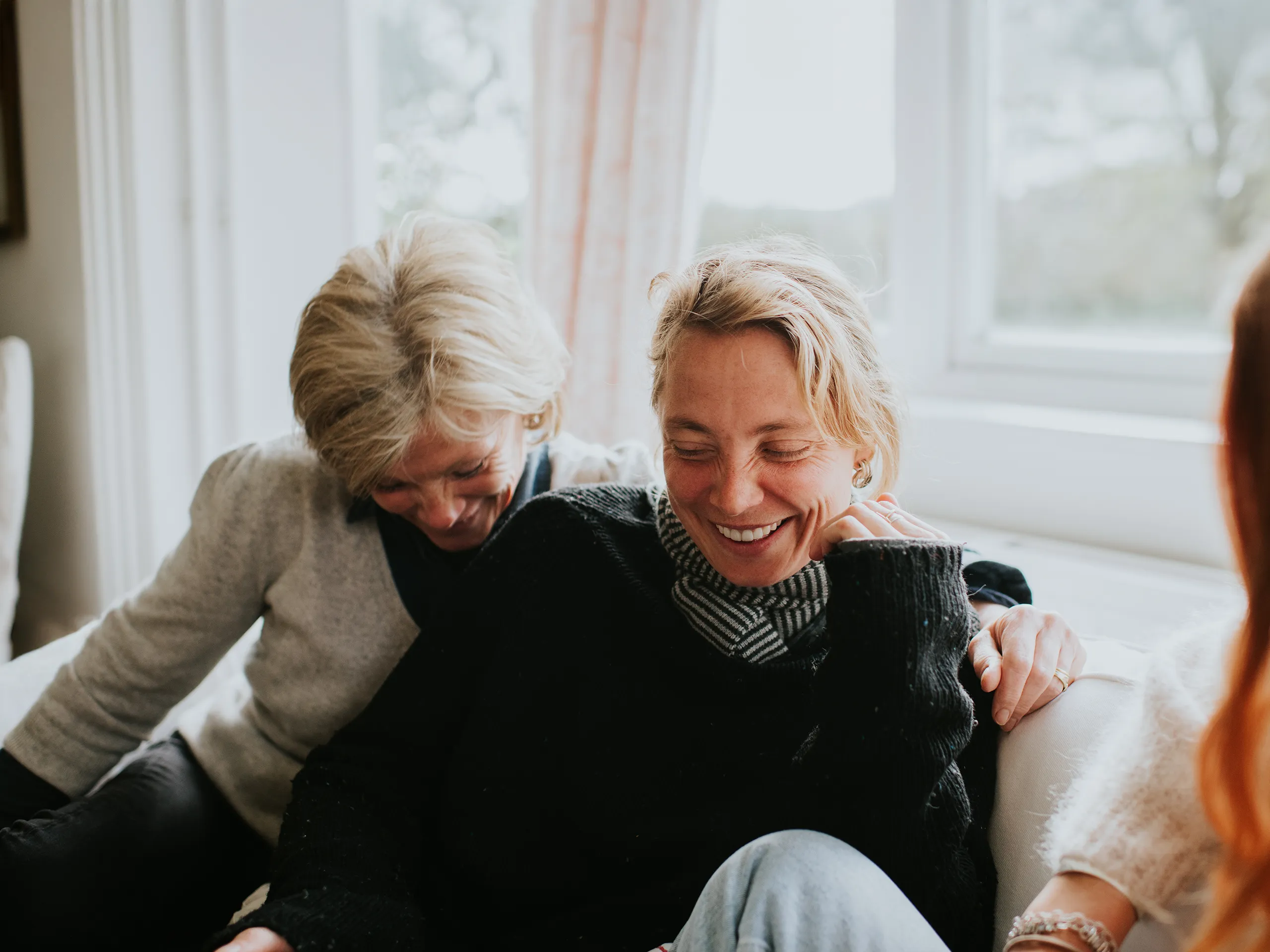 Two women laugh together on a sofa in a cozy atmosphere.