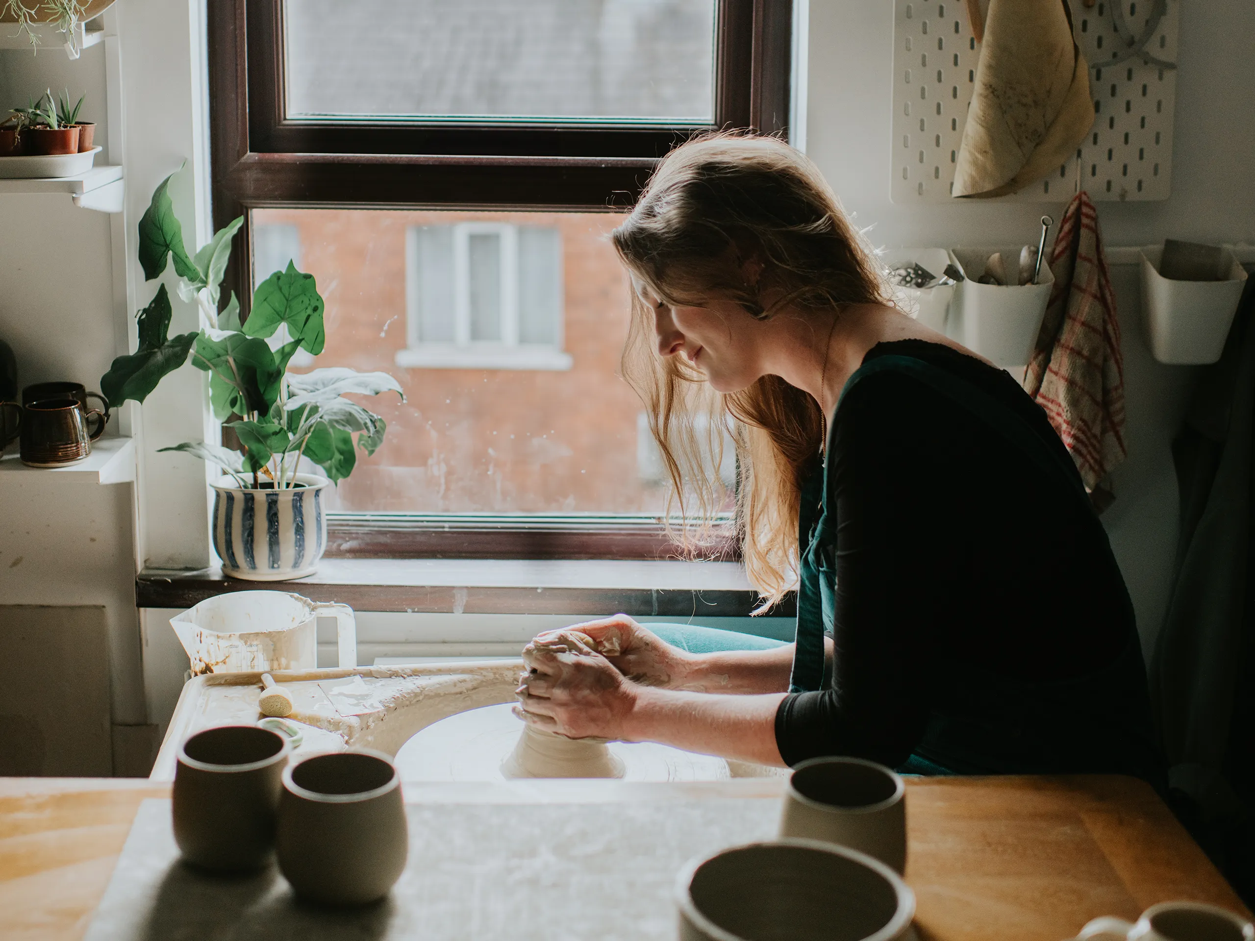 Woman works at a pottery wheel in her studio in front of a window.